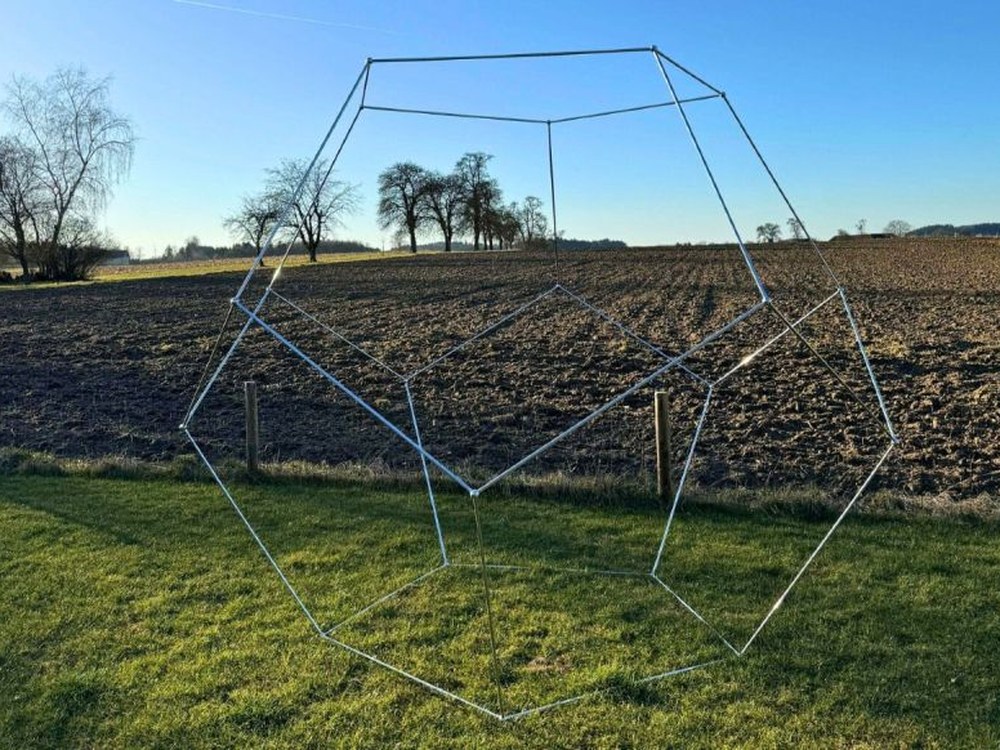 Großes Dodekaeder-Gestell aus Metallstangen auf einer Wiese vor einem Feld